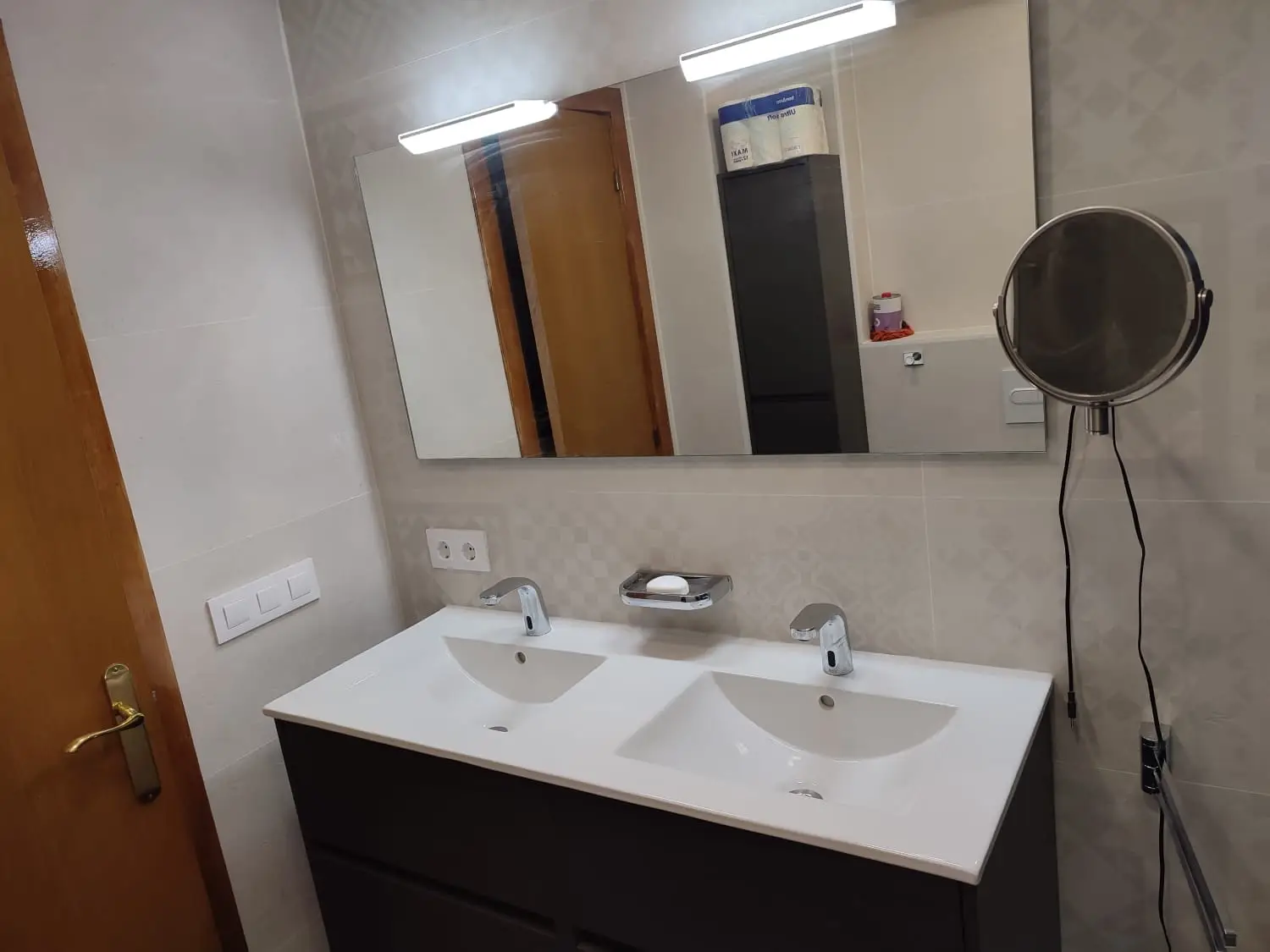 Baño con lavabo doble sobre mueble gris oscuro, espejo con iluminación y revestimiento claro